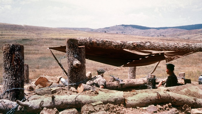 In einer trockenen Landschaft steht ein Unterstand aus Holz und Tüchern. Darunter stehen zwei deutsche Legionäre und blicken in die Landschaft.