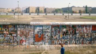 1980er: Ein Mann läuft auf der Westseite der Berliner Mauer entlang, dahinter sieht man den Todesstreifen und hinter einer weiteren Mauer einige Gebäude in Ost-Berlin