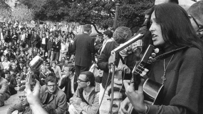 Rechts im Vordergrund steht eine Sängerin mit Gitarre. Im Hintergrund sind viele sitzende und stehende Menschen zu sehen.