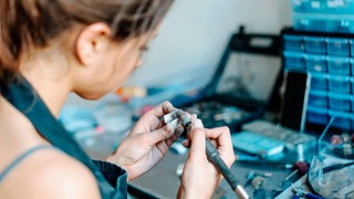 Eine Frau sitzt an einem Tisch, in der Hand hält sie einen elektrischen Schleifer, mit dem sie ein Stück Schmuck bearbeitet.