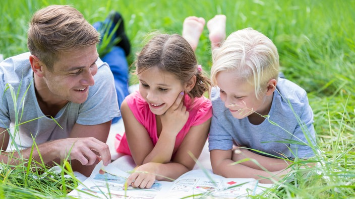 Ein  Mann liest mit zwei Kindern auf einer Wiese ein Heft