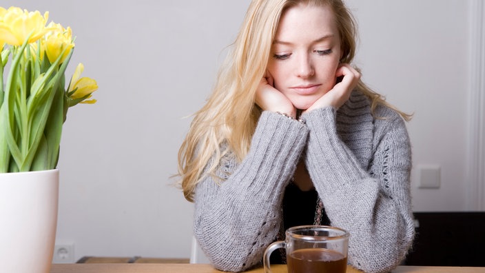 Eine junge Frau sitzt am Tisch und schaut traurig auf ihre Teetasse.