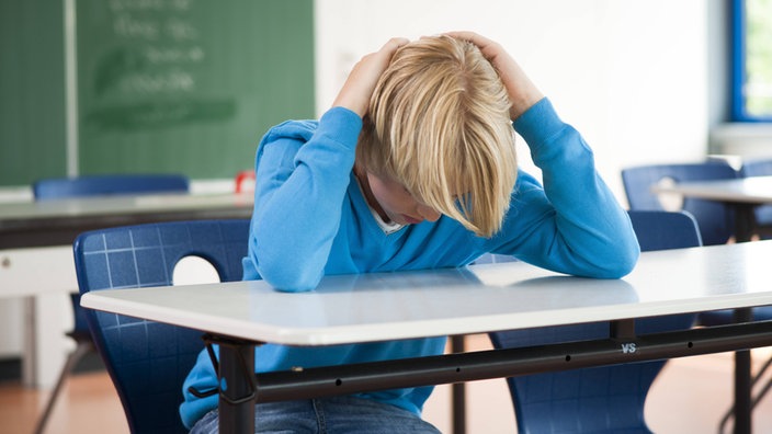 Blonder Junge sitzt in einem Klassenzimmer. Er hat die Arme und Hände über seinem Kopf zusammen geschlagen.