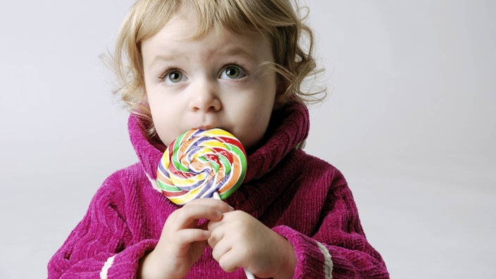 Das Foto zeigt ein kleines Mädchen, das einen großen, bunten Lolli in den Händen hält