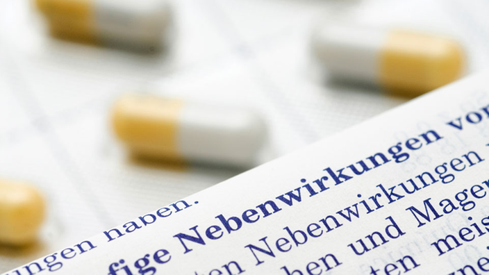 Nahaufnahme eines Beipackzettels mit dem Wort "Nebenwirkung" im Vordergrund und dahinter gelb-weiße Kapseln.