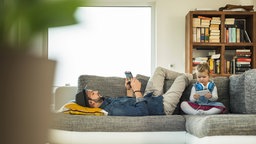 Ein Vater und ein Kind sitzen auf einem Sofa und schauen auf Tablet und Smartphone.