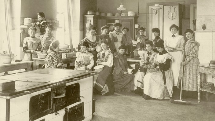 Schwarzweiß-Foto einer Gruppe von Frauen, die neben einem großen Herd sitzen und stehen