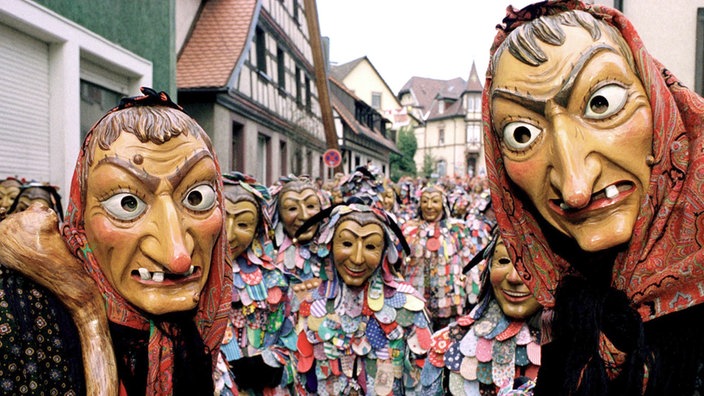 Zwei Narren mit Hexenmasken rahmen weitere bunt gekleidete Narren ein.