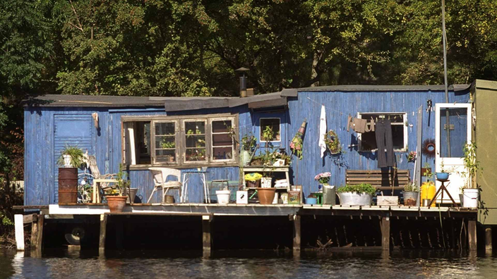 Blau gestrichene Holzbaracken stehen auf Pfählen im Wasser. Die Eingänge schmücken zahlreiche Blumentöpfe, Tische und Stühle. Hinter den Behausungen stehen dicht an dicht hohe Bäume.