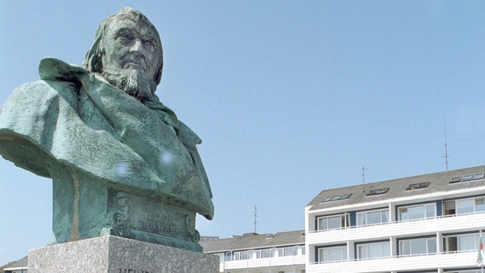 Links im Bild ist das Fallersleben-Denkmal zu sehen, das von Touristen betrachtet wird. Im Hintergrund stehen Mehrfamilienhäuser.