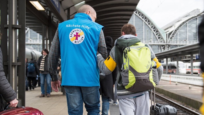 Ein Mitarbeiter der Bahnhofsmission  begleitet einen sehbehinderten Fahrgast am Bahnsteig.