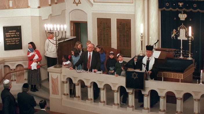 Gottesdienst in einer Synagoge. Ein Mann steht hinter einem Pult und spricht zur Gemeinde