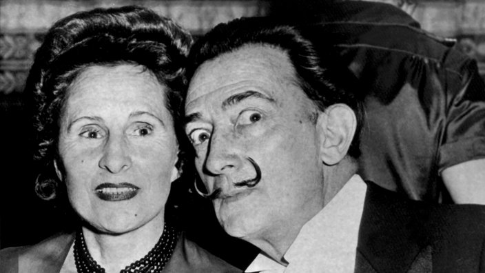 Schwarzweiß-Foto von Gala und Dalí bei einem Essen. Dalí beugt sich zu ihr hin und verdreht die Augen, sie lächelt in die Kamera.