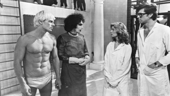 Eine Schwarzweiß-Filmszene aus dem Kinostreifen 'Rocky Horror Picture Show'. Frank'n'Furter präsentiert Brad und Janet seine neu erschaffene Mensch-Kreation.