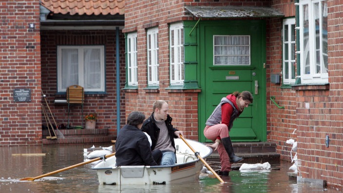 Zwei Männer rudern mit einem Boot durch das Dorf Hitzacker. Eine Frau in Gummistiefeln verlässt das Boot nahe einem Haus.