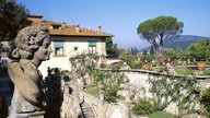 Ein Garten mit vielen Mauern und Blumentöpfen. Im Hintergrund steht eine alte Villa. Links im Vordergrund ist eine Frauenbüste zu sehen.