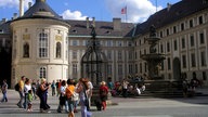 Das Foto zeigt ein helles herrschaftliches Gebäude. Auf dem Hof flanieren sommerlich gekleidete Touristen.