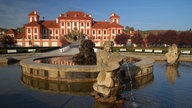 Im Vordergrund ist ein üppiger runder Brunnen zu sehen, im Hintergrund ein weiß-rotes Gebäude.