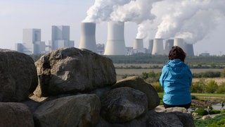 Eine Frau sitzt auf einem großen Stein, im Hintergrund das Braunkohlenkraftwerk Boxberg