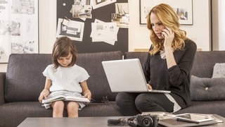 Eine Frau mit Laptop und Handy sitzt auf der Couch, daneben ihre kleine Tochter mit einem Buch