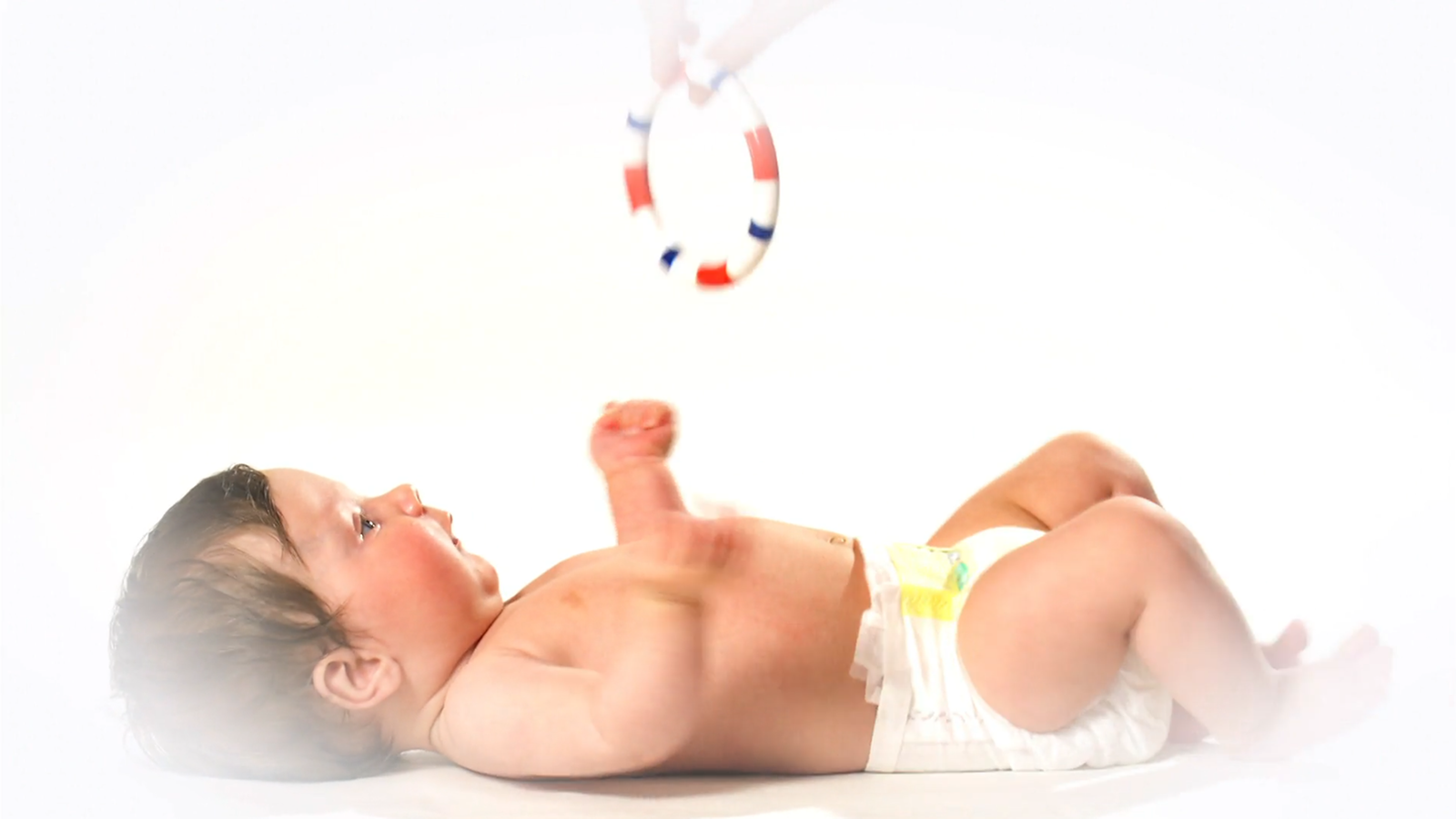 Ein Baby liegt auf einer Decke, eine Hand hält ihm einen bunten Ring zum Spielen entgegen.